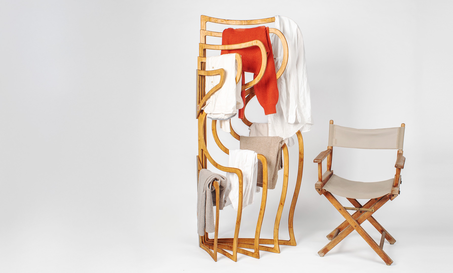 Relief, a wave-shaped wooden hanger, holding shirts and a red knit sweater, standing next to a director's chair, on a wide studio backdrop
